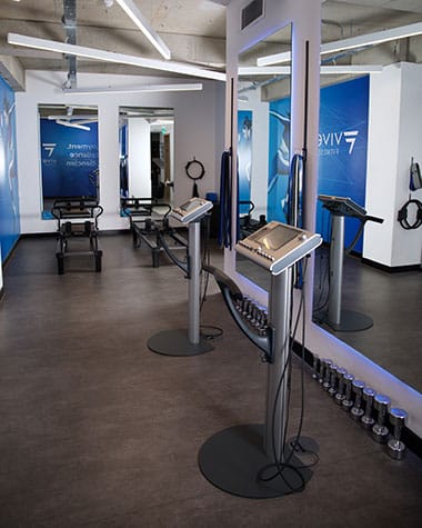 A bright studio with white brick walls and large mirrors, the studio floor featuring ems equipment and reformer pilates equipment.