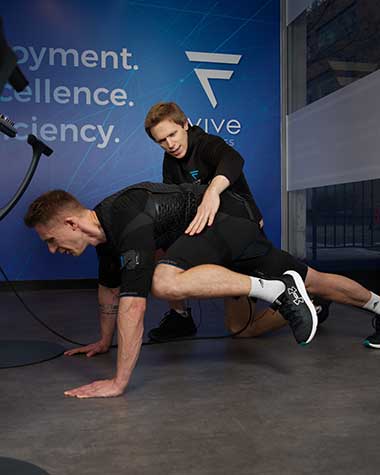 A client in an EMS suit performing an EMS stretch while receiving positive feedback from a personal trainer.