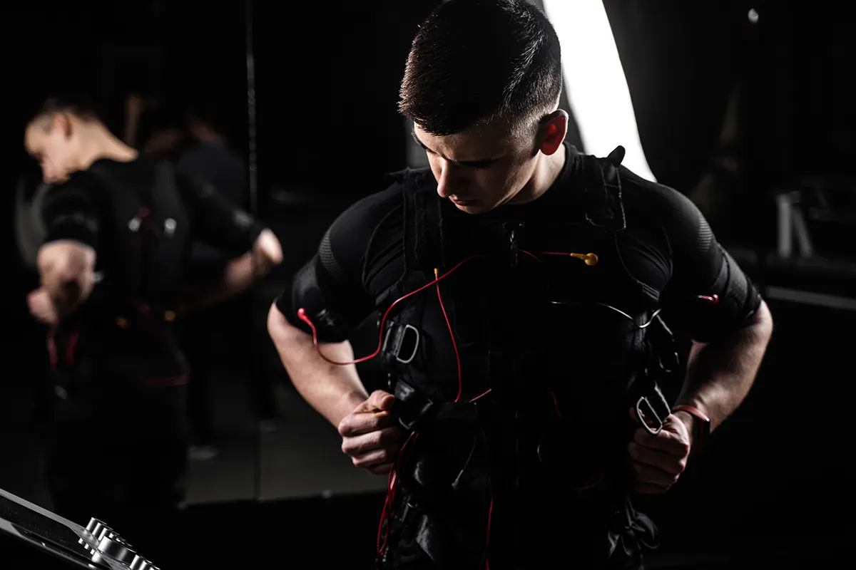 Man adjusting a black EMS (Electrical Muscle Stimulation) training suit with red electrode wires in a dimly lit studio environment.