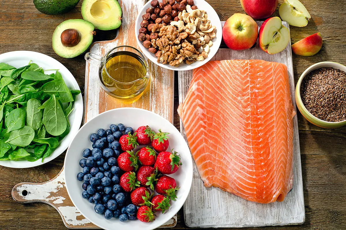 Flat lay overhead view of healthy whole foods including a raw salmon fillet, fresh berries, spinach, avocados, mixed nuts, apples, olive oil, and seeds.