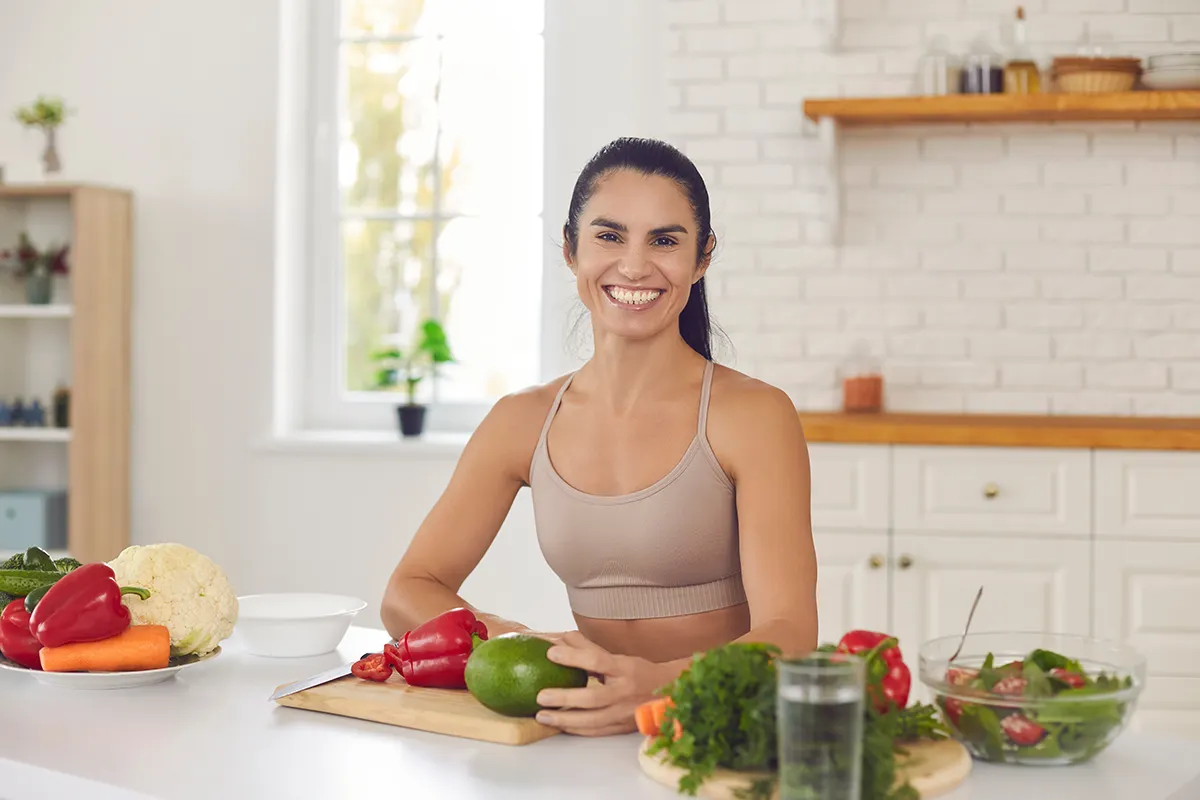 Smiling woman in activewear preparing fresh vegetables like avocado and bell peppers in a bright kitchen for a healthy meal.