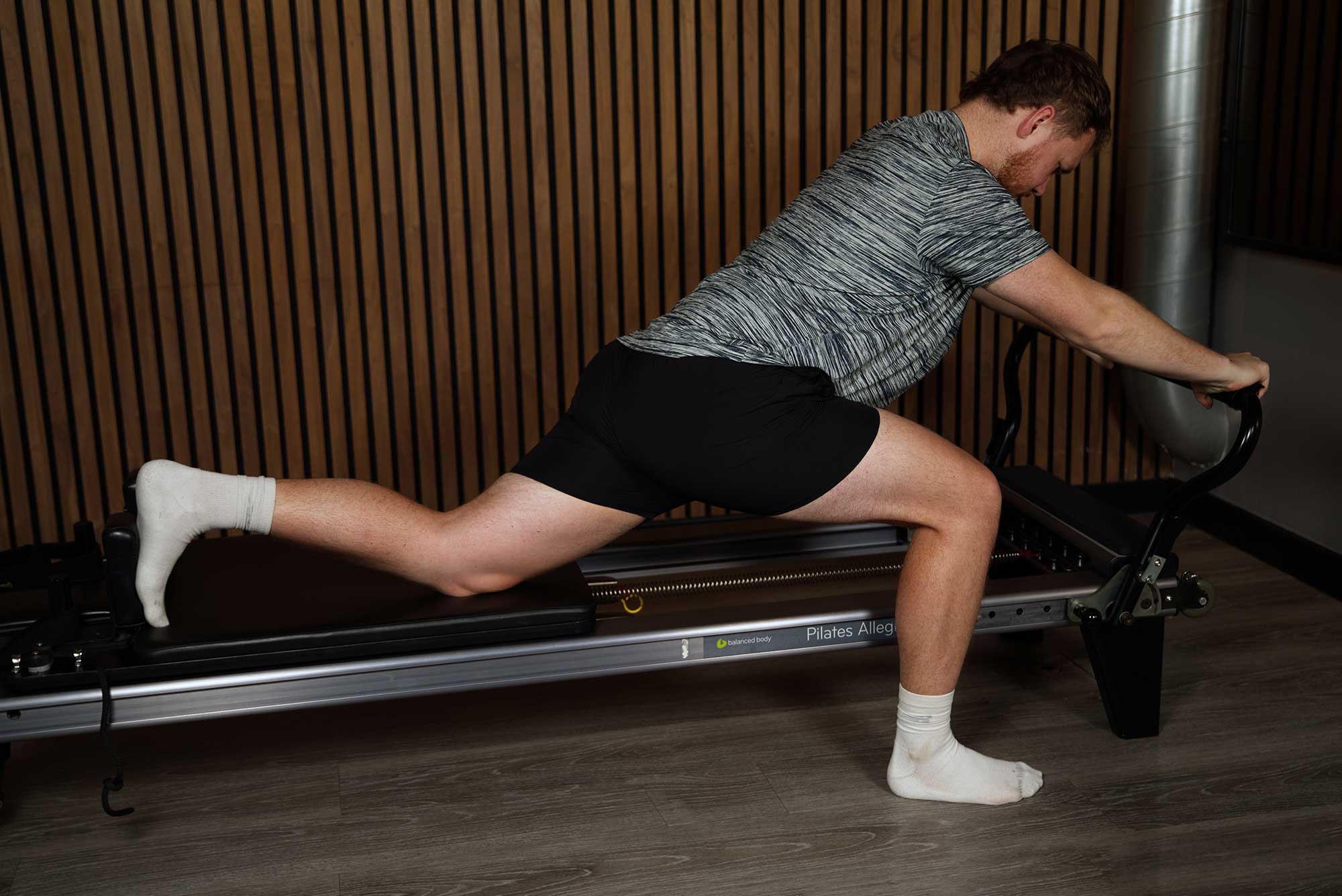 A male client performing a core-focused plank exercise on a reformer, guided by a personal trainer.
