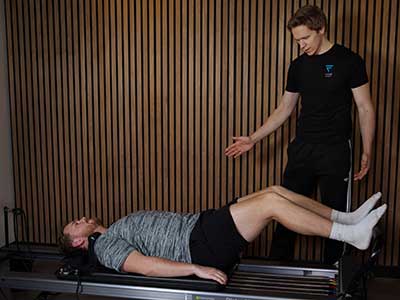 A personal trainer from Vive Fitness monitoring a client using a reformer Pilates machine in front of a wooden-slat wall.