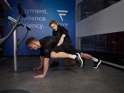 A client in an EMS suit performing an EMS stretch while receiving positive feedback from a personal trainer.